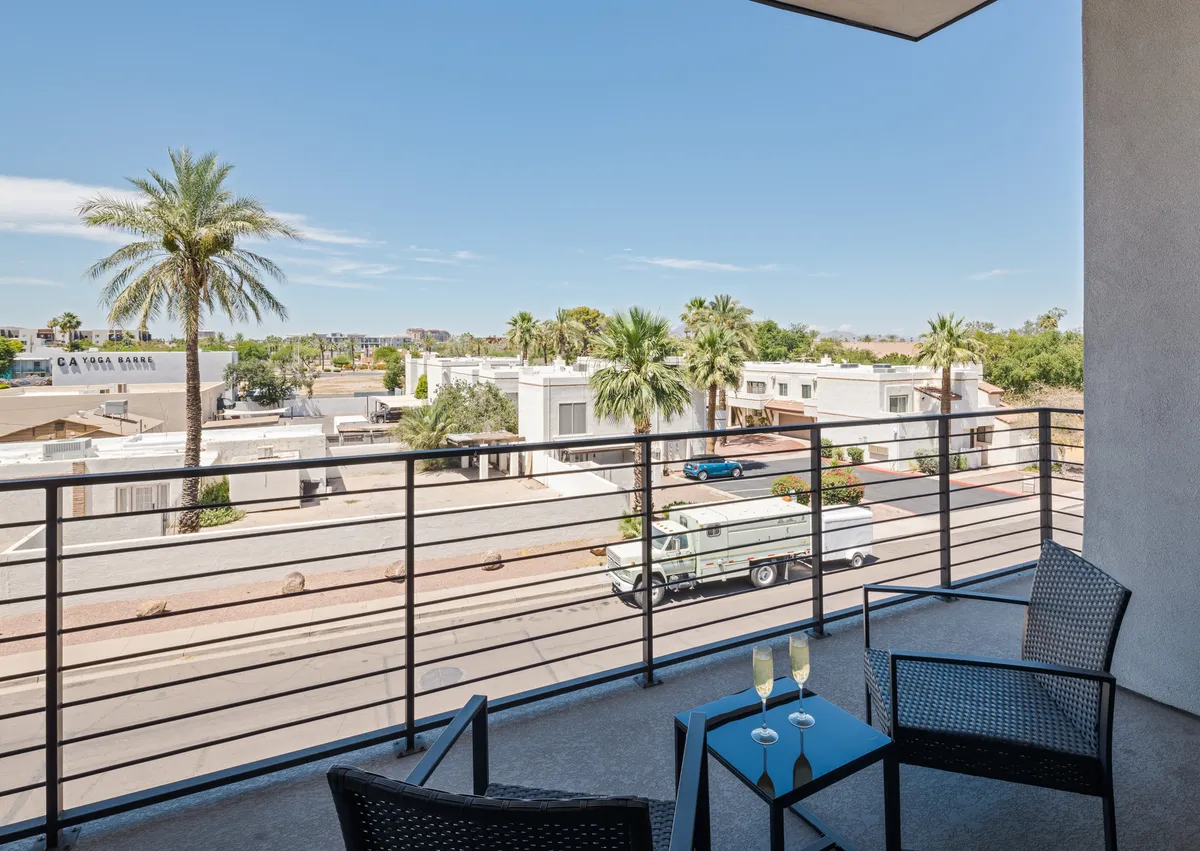 This exterior image highlights a balcony with seating, palm trees in the background, and a sunny blue sky. The setting evokes a laid-back, social atmosphere ideal for enjoying coffee or relaxing outdoors with your pet.