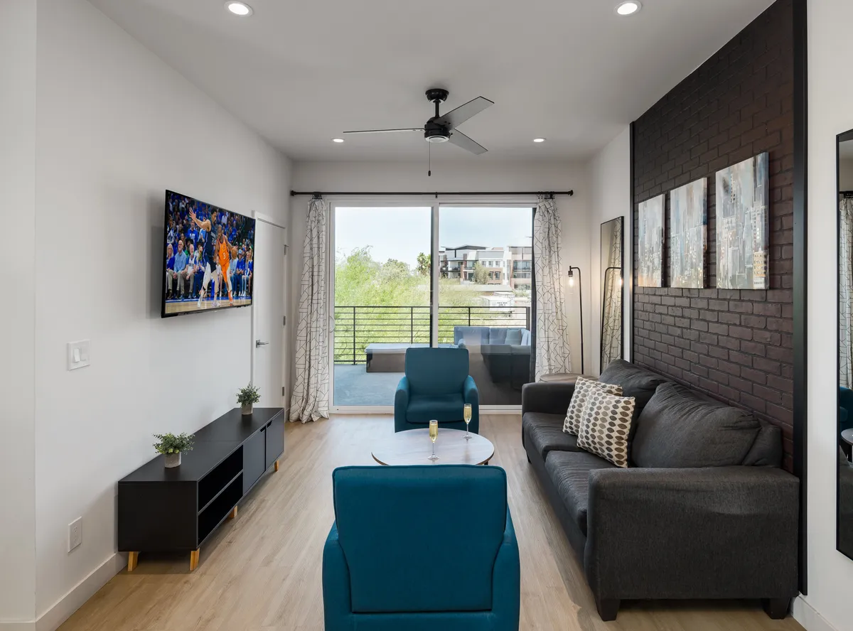 The photo features a living space with a sleek TV displaying a sports event, modern furniture, and a large sliding glass door leading to a private balcony. The room is bathed in natural light and designed for both lounging and social interaction.