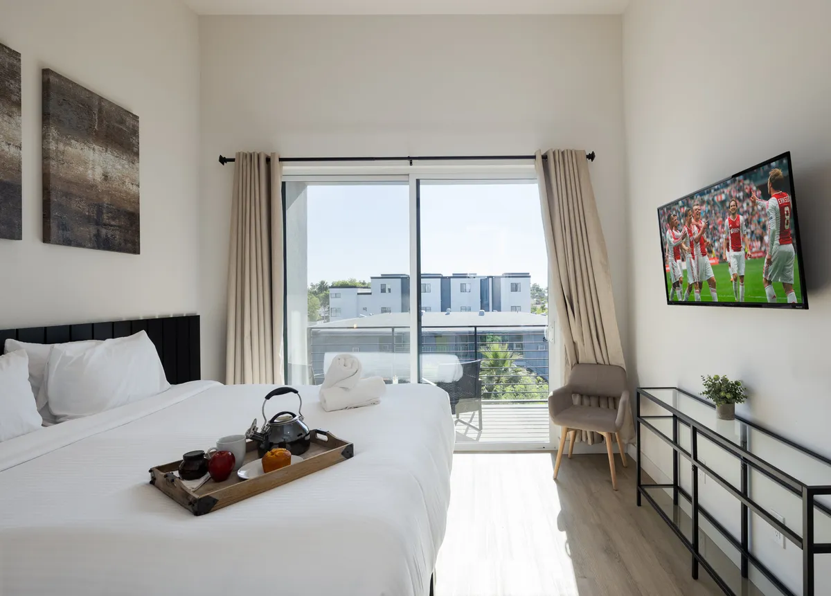 A comfortably appointed bedroom featuring a bed with crisp white linens, a wall-mounted Smart TV, a tray with breakfast items, and a sliding glass door showcasing a balcony view. The overall mood is casual and inviting.