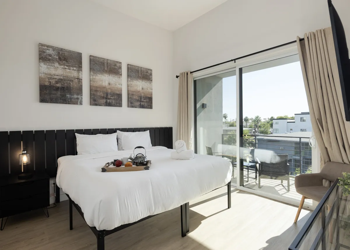 A serene bedroom with a plush bed topped with white linens, a breakfast tray, and a large sliding glass door leading to a private balcony. Abstract art hangs on the wall, and the room is bathed in natural light, emphasizing tranquility and comfort.