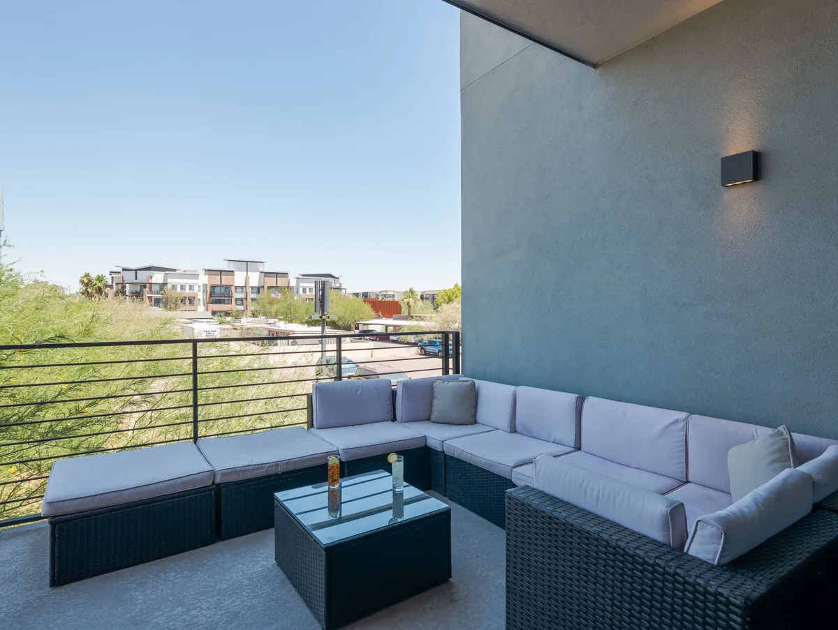 An inviting private balcony sets the scene, complete with a sectional sofa, glass coffee table, and a scenic view of neighboring buildings. The setting is perfect for relaxing, entertaining, or enjoying the cityscape at sunset.