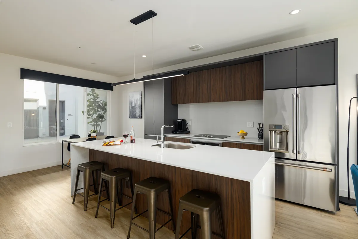 A welcoming kitchen scene featuring a spacious island with bar stools, stainless steel appliances, and abundant natural light streaming in from a window. The setup is ideal for both cooking and casual dining, inviting guests to gather around the island.