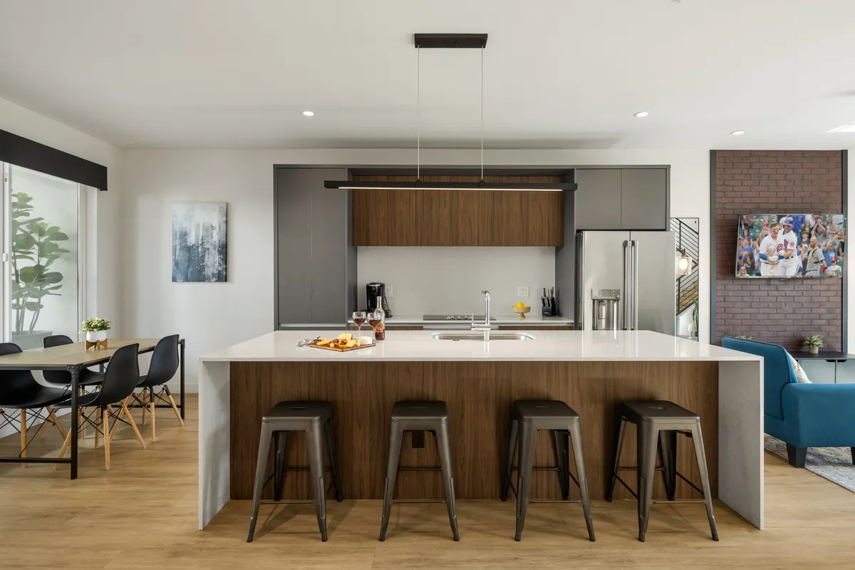A contemporary kitchen and dining area with a sleek island, bar stools, visible refrigerator, and a dedicated dining space with modern artwork. The layout supports both food preparation and shared meals, aligning with the theme of communal dining and culinary gatherings.