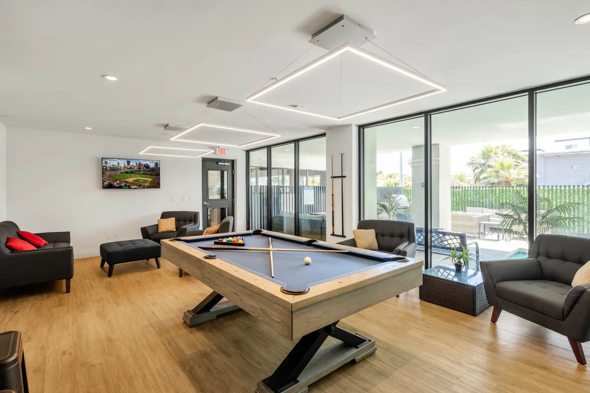 A vibrant communal lounge featuring a pool table at the center, surrounded by comfortable sofas, with large windows letting in abundant natural light. A television is mounted on the wall for group sports viewing or movie nights.