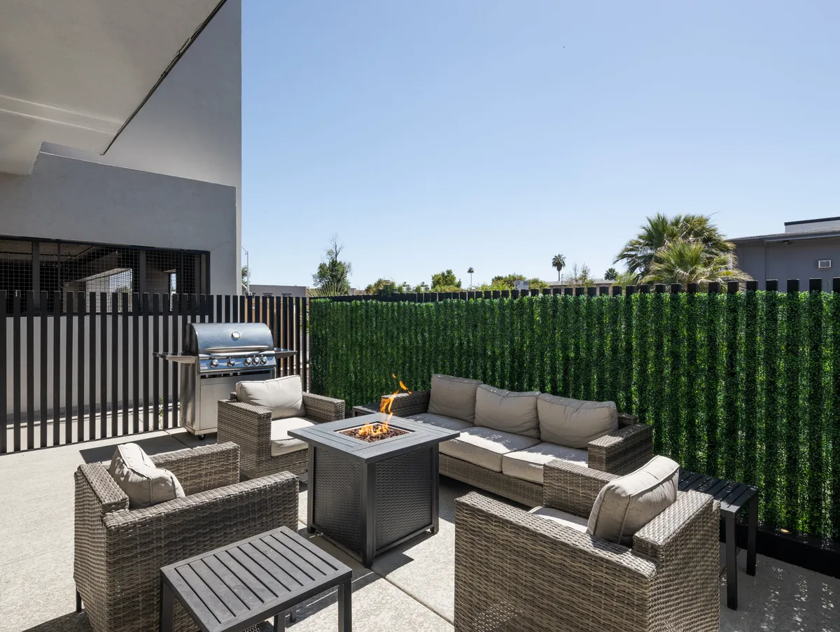 An outdoor space designed for togetherness, featuring a fire pit surrounded by seating, a BBQ grill in the background, and faux greenery for ambiance. The setting is perfect for grilling, gathering around the fire, and relaxing under the open sky.